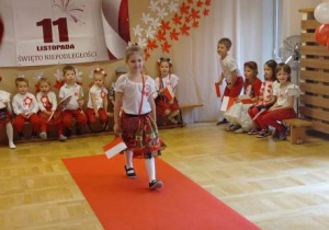Dziewczynka prezentuje na czerwonym dywanie strój krakowianki, w tle grupa dzieci