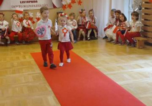 Dziewczynka i chłopiec na czerwonym dywanie prezentują strój, w tle grupa dzieci