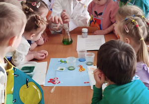 Dzieci przy stoliku, patrzące na panią, która wlewa do zlewki zielony płyn