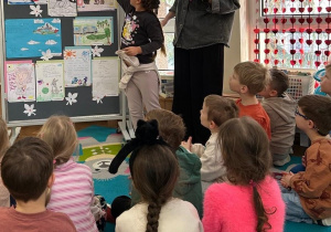 Grupa dzieci patrzy na dziewczynką stojącą obok nauczyciela i pokazującą swoją pracę na tablicy